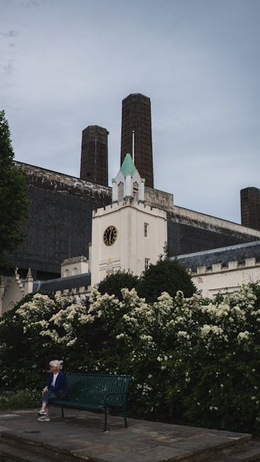 A person seated on a green park bench in front of a white church with small green spires and a clock on its facade, surrounded by lush flowering bushes with white blossoms. Behind the church, there are tall industrial-style brick chimney stacks under a cloudy sky, indicating an urban setting. The scene appears to be part of a residential or public area near Battersea, with the church positioned close to the street. This image could relate to house removals or furniture transport in the Battersea area, demonstrating the typical environment where a professional removals service like Man and a Van Battersea might operate during home relocation or moving logistics, focusing on the urban surroundings and street-level activity.