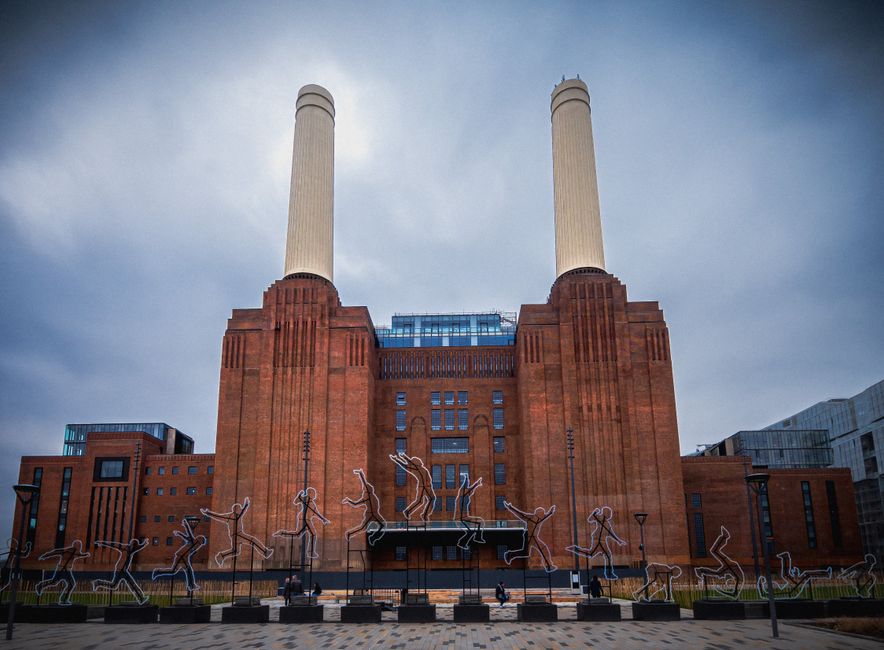An upward perspective of an industrial brick building during daytime, showing two large, cylindrical, cream-colored chimneys extending vertically from the structure's flat roof. The facade of the building features multiple rectangular windows with black metal grids, arranged in a grid pattern across the brick surface. The building's exterior includes vertical brick pilasters and a parapet, with some windows covered by metal shutters. The sky above is partly cloudy with patches of blue visible through the clouds. This scene illustrates the type of structure involved in house removals or furniture transport, highlighting the importance of careful logistical planning when relocating items from such industrial environments, as managed by services like Man and a Van Battersea.
