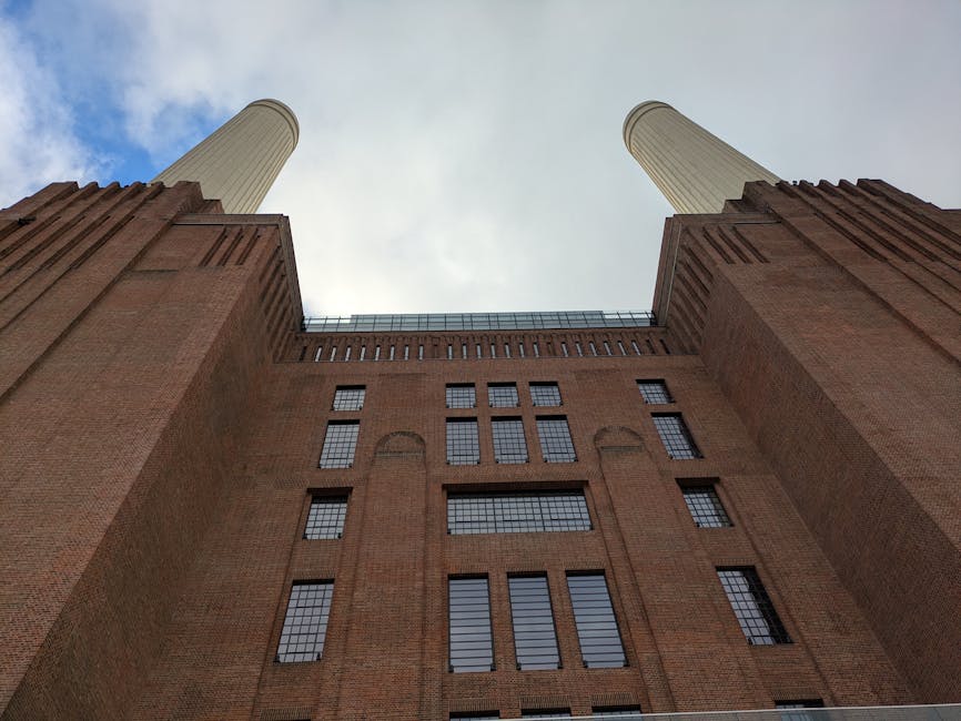 Ground-level view of a large brick building with multiple rectangular windows, characteristic of an industrial or warehouse structure, flanked by two tall, cylindrical white chimneys extending upward into the sky. The building's facade features a series of window arrangements with some panes covered by metal grilles. The sky above is partly cloudy with patches of blue visible. This scene may relate to a home relocation or furniture transport project by Man and a Van Battersea, highlighting the building's architectural environment associated with moving and packing processes.