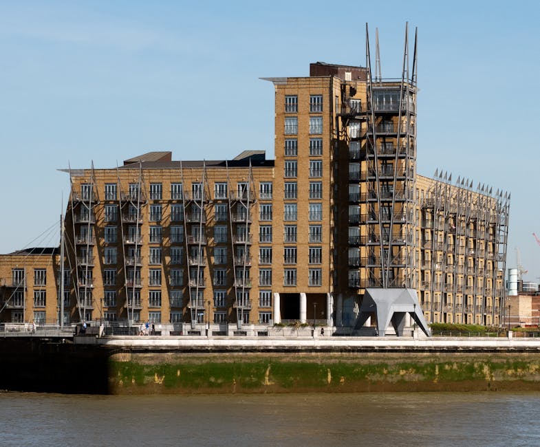 Photograph of a modern multi-storey residential building located on the riverside, with scaffolding and construction equipment visible around the exterior indicating ongoing development or renovation. The building features numerous windows and small balconies, with the structure primarily constructed from brick. In front of the building, there is a paved area or roadway where a man and a van are likely working, as part of a home relocation or furniture transport process. The van is parked close to the building, on the pavement, and is presumed to be loaded or unloading household items. The ground level includes an open area possibly used for access or staging during the moving activities. The overall scene captures the urban environment and the logistics involved in house removals, with the presence of construction elements suggesting a growing or redeveloped estate, fitting for a destination like Winstanley Estate in Battersea, SW11, serviced by Man and a Van Battersea.